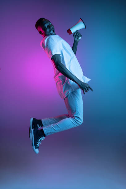 istock Young smiling jumping african man in neon light on gradient background. Concept of emotions, action, facial expression. 1325418439