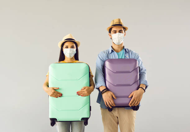 istock Young couple in face masks holding suitcases standing on light gray studio background 1325420586