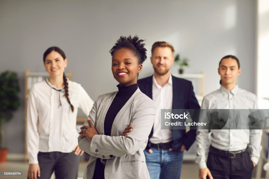 African-American businesswoman standing arms folded and smiling with team of workers behind Happy aspiring black business woman in suit smiling at camera standing arms folded with team of workers behind. Portrait of ambitious female leader, executive, bank staff manager. Blurred background Business Stock Photo