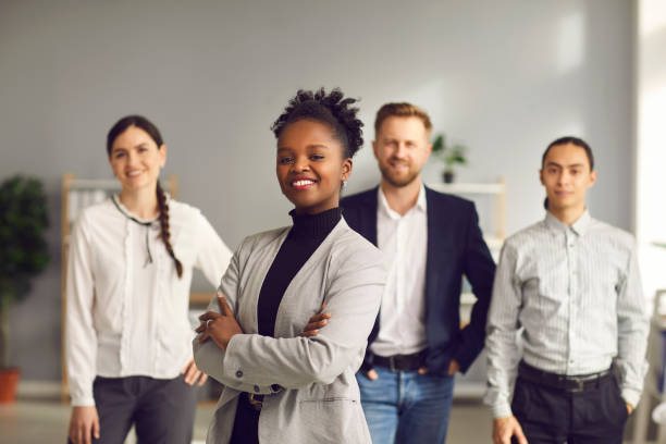 African-American businesswoman standing arms folded and smiling with team of workers behind Happy aspiring black business woman in suit smiling at camera standing arms folded with team of workers behind. Portrait of ambitious female leader, executive, bank staff manager. Blurred background founder stock pictures, royalty-free photos & images