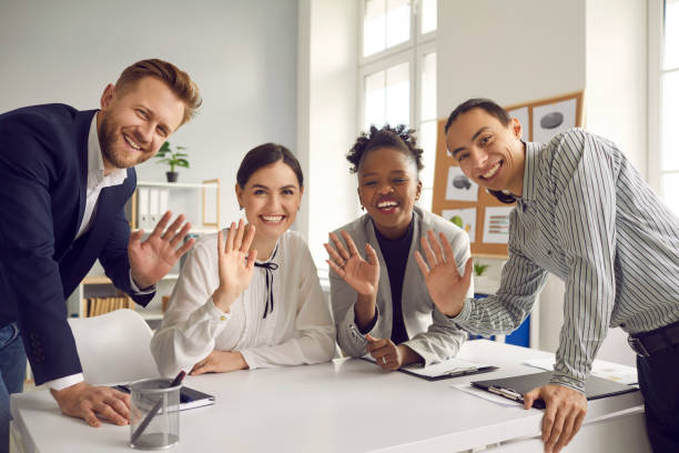 Team of business people waving hello at camera greeting international coworkers in online meeting Team of happy young diverse business people sitting and standing around office table, smiling, looking at camera and waving hello, greeting international coworkers in remote work meeting via Internet greeting stock pictures, royalty-free photos & images