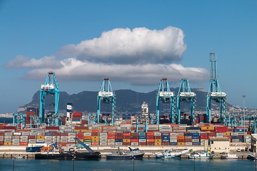 Algeciras, Cadiz, Spain - June 24, 2021: Ships, containers and cranes in the commercial port of Algeciras, with the rock of Gibraltar in the background with a large cloud above