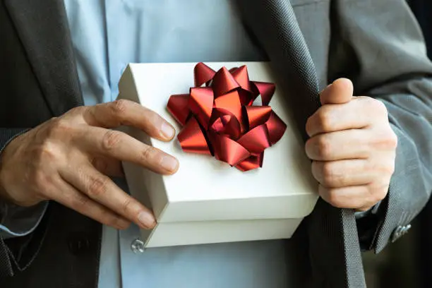 Midsection of an unrecognizable businessman wearing a suit who is about to give a gift box out of his jacket. The white gift box is wrapped with red ribbon.