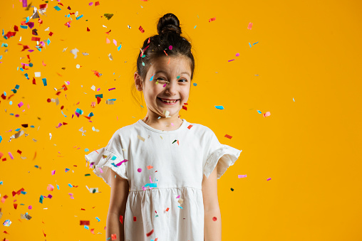 Caucasian girl is standing under confetti falling down and is looking at camera in front of yellow background.