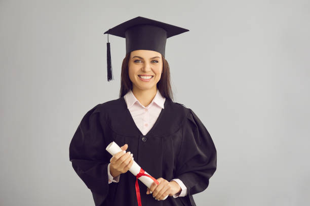 female graduate standing on a gray background with a diploma in her hands and smiling. - grad portrait imagens e fotografias de stock