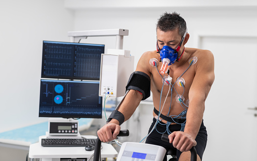 Doctors observing the progress of a cardiopulmonary stress test taken by the male athlete riding a bicycle ergometer. Young adult man having a VO2 test with a VO2 mask on his face, electrocardiogram pads attached, computer recording, indoor bicycle.