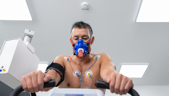 Doctors observing the progress of a cardiopulmonary stress test taken by the male athlete riding a bicycle ergometer. Young adult man having a VO2 test with a VO2 mask on his face, electrocardiogram pads attached, computer recording, indoor bicycle.