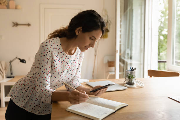 istock Smiling woman writing taking notes in notebook, holding smartphone 1325452723