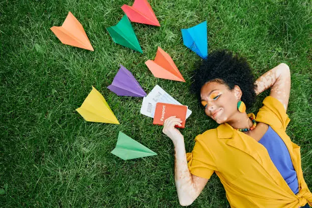Traveling concept. Multi-ethnic woman with air tickets and international passport in hand dreams to vacations lying on green grass around colored paper airplanes