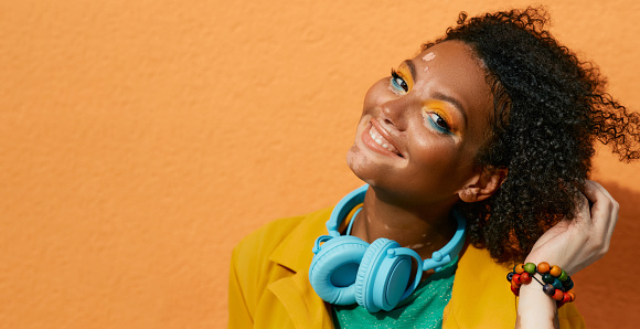 Portrait of a laughing woman with vitiligo wearing in blue headphones and colorful clothes on background of orange city wall. Bright lifestyle