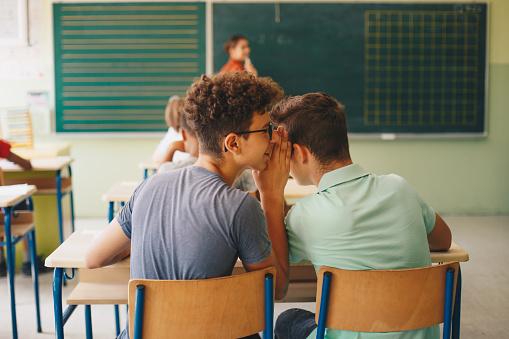 Two teenage boys are sitting at school desk and whispering during class while teacher is questioning class. They are sitting next to each other and talking. Selective focus.