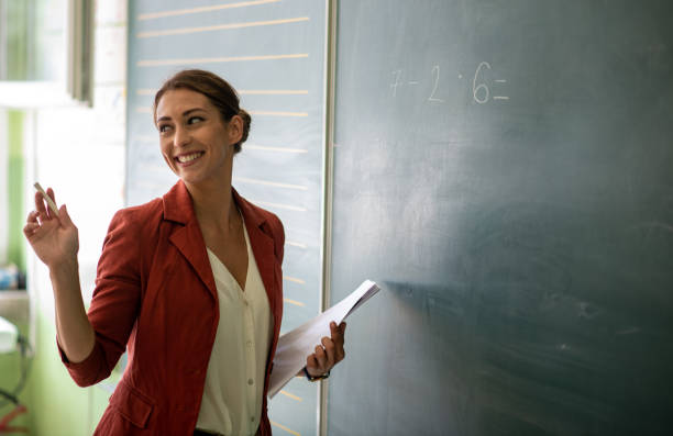 Young smiling female teacher is standing in front of a blackboard and explaining to kids mathematical operations. She has white chalk in one hand and papers in other hands.