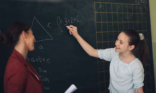 Teacher is questioning young girl in front of a class. School girl is solving math problem and calculating circumference of a triangle. She is happy because she had chance to show her knowledge in front of her friends.