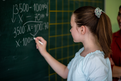 Schoolgirl with ponytail volunteered to go in front of her class and solve equation. She loves mathematics and school and she is glad to show her knowledge in front of her friends from class. Back to school concept.