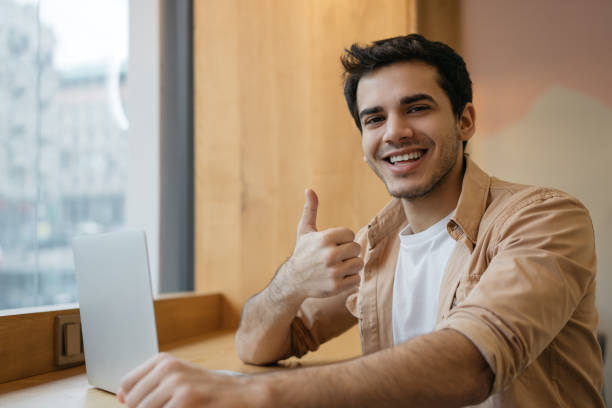 istock Smiling Indian man using laptop computer, showing thumb up, looking at camera working from home. Happy asian student studying, learning language, watching online training courses 1325465864