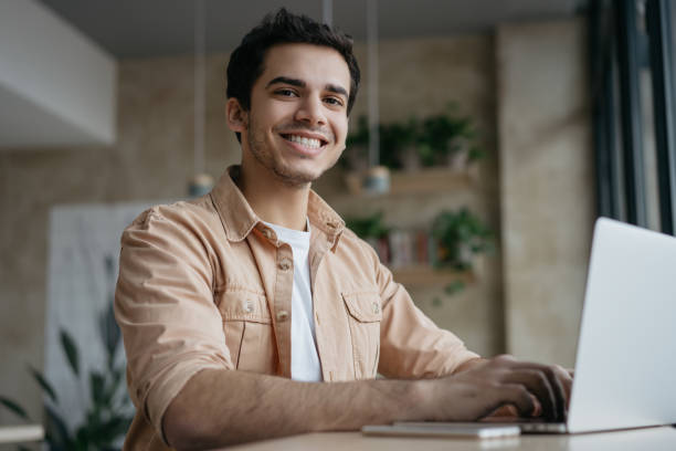 heureux étudiant indien étudiant, apprenant la langue. homme d’affaires métis utilisant un ordinateur portable, tapant sur le clavier, regardant l’appareil photo et souriant. projet indépendant de rédacteur travaillant en collaboration - technology typing male beauty asian and indian ethnicities photos et images de collection