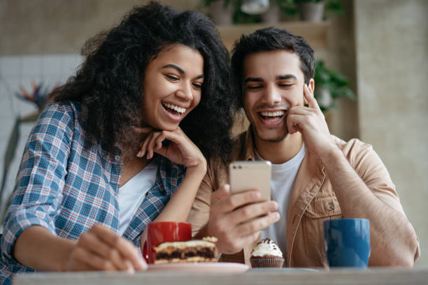 istock Happy couple using mobile phone and internet, watching movie together, communication, laughing, sitting in cafe. Positive hipster friends shopping online, looking at digital screen 1325465986
