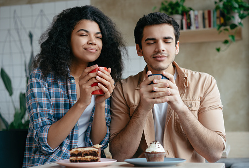 Multiracial friends drinking coffee together, relaxing in cafe. Beautiful African American woman and attractive Indian man holding cups of tea, enjoying life. Coffee break, perfect morning concept