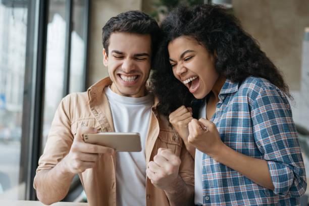 amigos emocionados usando el teléfono inteligente, jugando videojuegos. feliz éxito de celebración de la pareja emocional. retrato de los ganadores de la lotería en línea muy contentos mirando la pantalla digital. concepto de apuestas deportivas - couple young women casual excitement fotografías e imágenes de stock