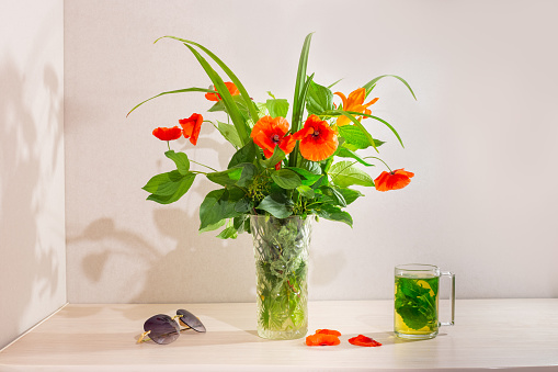 A bouquet of wildflowers in a vase on the table, next to a mug with mint leaves and sunglasses. Atmospheric summer mood.