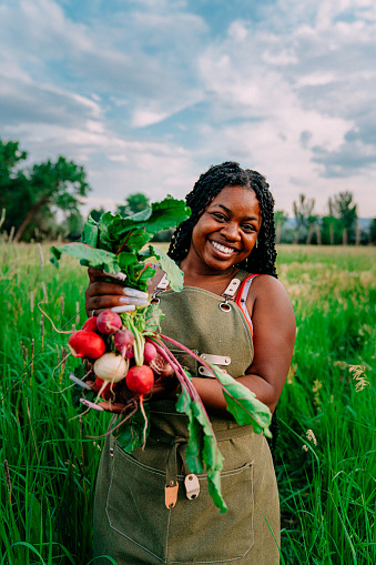 Portrait of a Happy Young Female African American Farmer Girl Standing in a Green Wheat Grass Field Outdoors Holding Delicious Fresh Produce at a Local Small Business Farm-to-Table Supplier in Colorado