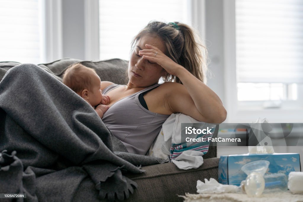 Postpartum stress A middle aged mother resting on her couch holds her baby in her arm and holds her other hand over her head with a tired and stressed expression. Mother Stock Photo