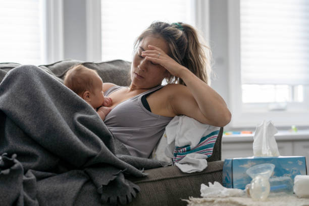 Postpartum stress A middle aged mother resting on her couch holds her baby in her arm and holds her other hand over her head with a tired and stressed expression. postpartum depression stock pictures, royalty-free photos & images