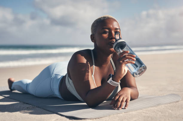 Shot of a sporty young woman drinking water while lying on her yoga mat at the beach