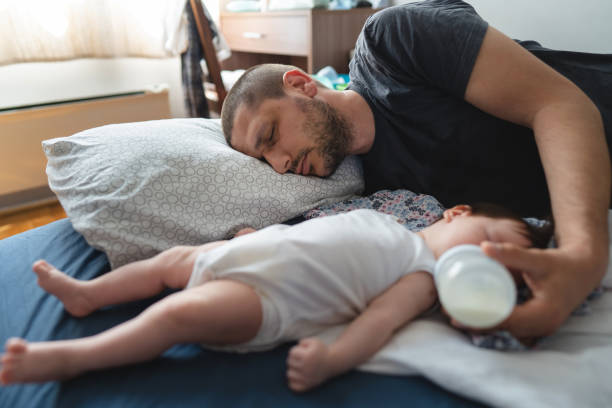 疲れ果てた男の父親は、幼児の親の概念選択的な焦点を与える哺乳瓶を保持しながら、自宅で寝ているベッドの上に彼の3ヶ月の赤ちゃんによって横たわっている - female nurse 写真 ストックフォトと画像