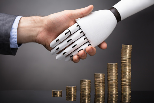 Businessperson And Robot Shaking Hands Over Stacked Golden Coins