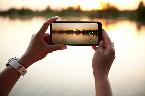 Close-up of female hands taking a photography of a beautiful summer sunset on the lakeshore background. Summer themes. Mobile phone in live view