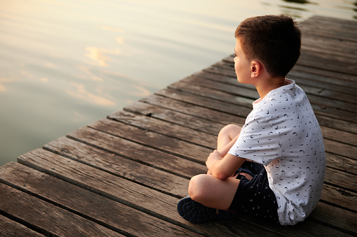 Serene schoolboy sitting on a pier and enjoying the tranquility of nature scene while looking at the lake landscape at sunset time