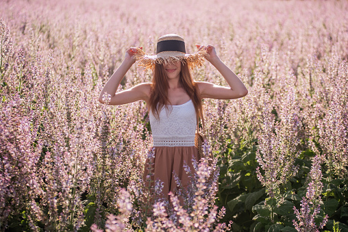 Young blonde woman in chiffon powdery jumpsuit against the background of blooming field of pink sage. Portrait of beautiful girl holding straw hat. Weekend walk outside the city. Agricultural texture