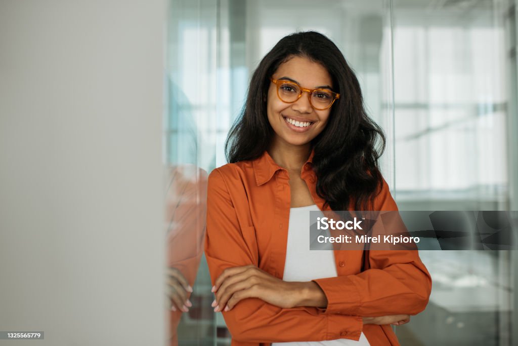 Mujer de negocios afroamericana sonriente que usa anteojos elegantes mirando la cámara de pie en la oficina moderna. Concepto exitoso de negocio y carrera - Foto de stock de Una sola mujer libre de derechos