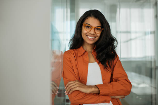 smiling african american business woman wearing stylish eyeglasses looking at camera standing in modern office. successful business and career concept - volwassen vrouwen stockfoto's en -beelden