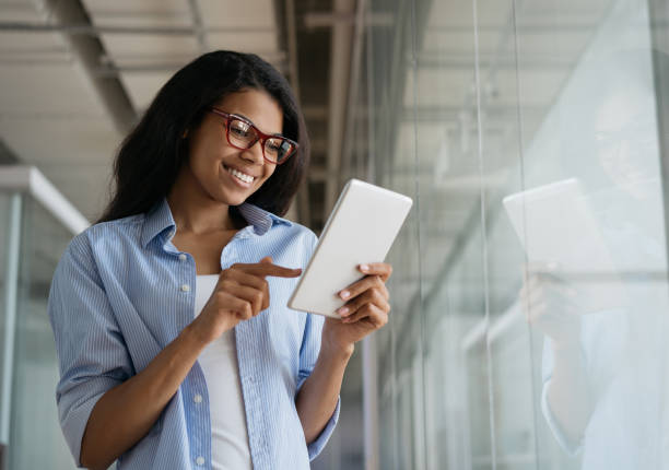 mulher afro-americana sorridente usando tablet digital trabalhando online em escritório moderno. estudos felizes do aluno, ensino a distância, conceito de educação - óculos de leitura - fotografias e filmes do acervo