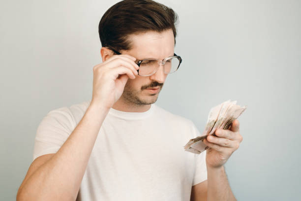 joven con grandes gafas mirando el dinero en sus manos, en el interior. conceptos salarios bajos, rentas que caen de la población, pobreza - low paid fotografías e imágenes de stock