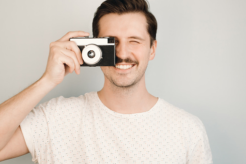 Young, smiling man holds vintage, retro camera and takes a photo indoors, close-up. Photographer hipster, film photo camera