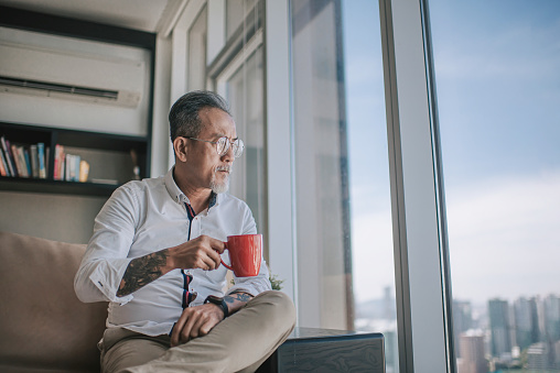 worried sad asian chinese senior man looking through window at living room sitting on sofa during day time