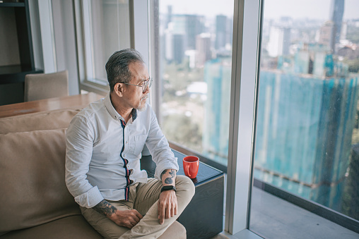 worried sad asian chinese senior man looking through window at living room sitting on sofa during day time