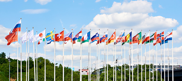 Group of flags of many different nations against blue sky and infront of a convention center.