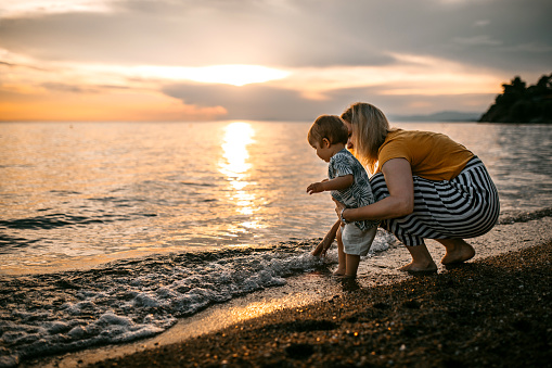 Mother and baby boy enjoy coastal walk and beautiful sunset at sand beach, splashing water and have fun