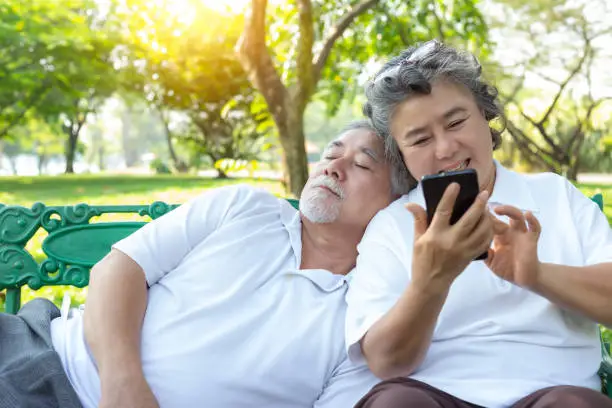 Photo of Happy Old woman using smart phone for chatting with family during old man or elderly husband sleeping on retirement wife shoulder at park Pensioner couple sitting on bench at public park and get relax