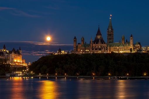 Canada’s Parliament Buildings and the historic Chateau Laurier hotel at night from across the river. Blue night sky with a full moon.