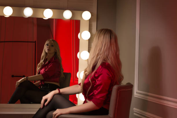 istock Luxurious blonde model wears red silk blouse, looking on her reflection in the big mirror with bulbs 1325610398