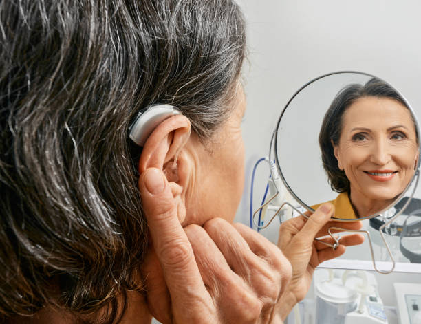 Mature woman with BTE hearing aid looks at herself in mirror and tries on hearing device, close-up. Hearing loss treatment Mature woman with BTE hearing aid looks at herself in mirror and tries on hearing device, close-up. Hearing loss treatment hearing aid photos stock pictures, royalty-free photos & images