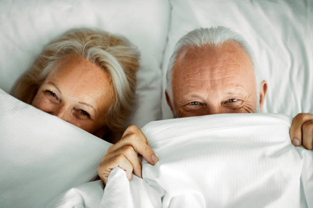 happy senior couple relaxing in bed hiding under sheets - sheet sleeping women bed imagens e fotografias de stock