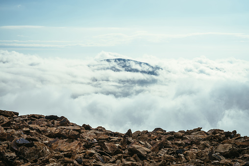 View from precipice edge over clouds to mountain vertex in thick clouds. Wonderful minimalist landscape with mountain top above dense low clouds. Scenic minimalism with mountain peak above cloudy sky.