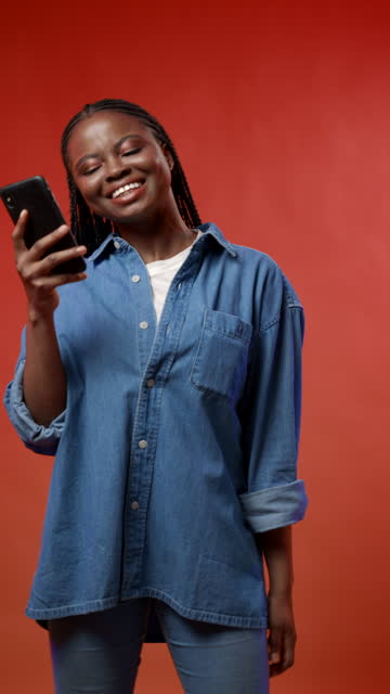 A vertical video of a cheerful attractive dark-skinned student buying something she wants very much in her smartphone with the credit card against the red background, she's satisfied that she finally bought what she wanted
