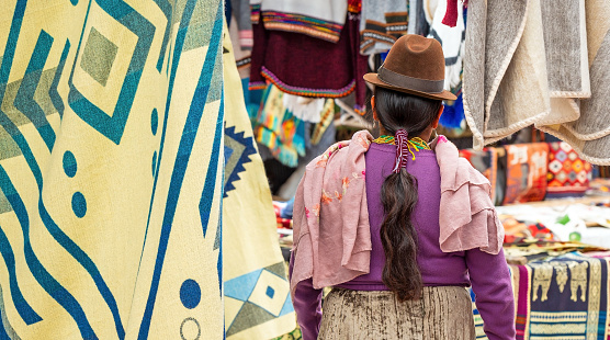 Indigenous Ecuadorian Otavalo woman in traditional clothing, hat and hairstyle on Otavalo local market with textile and fabric stalls, Ecuador.